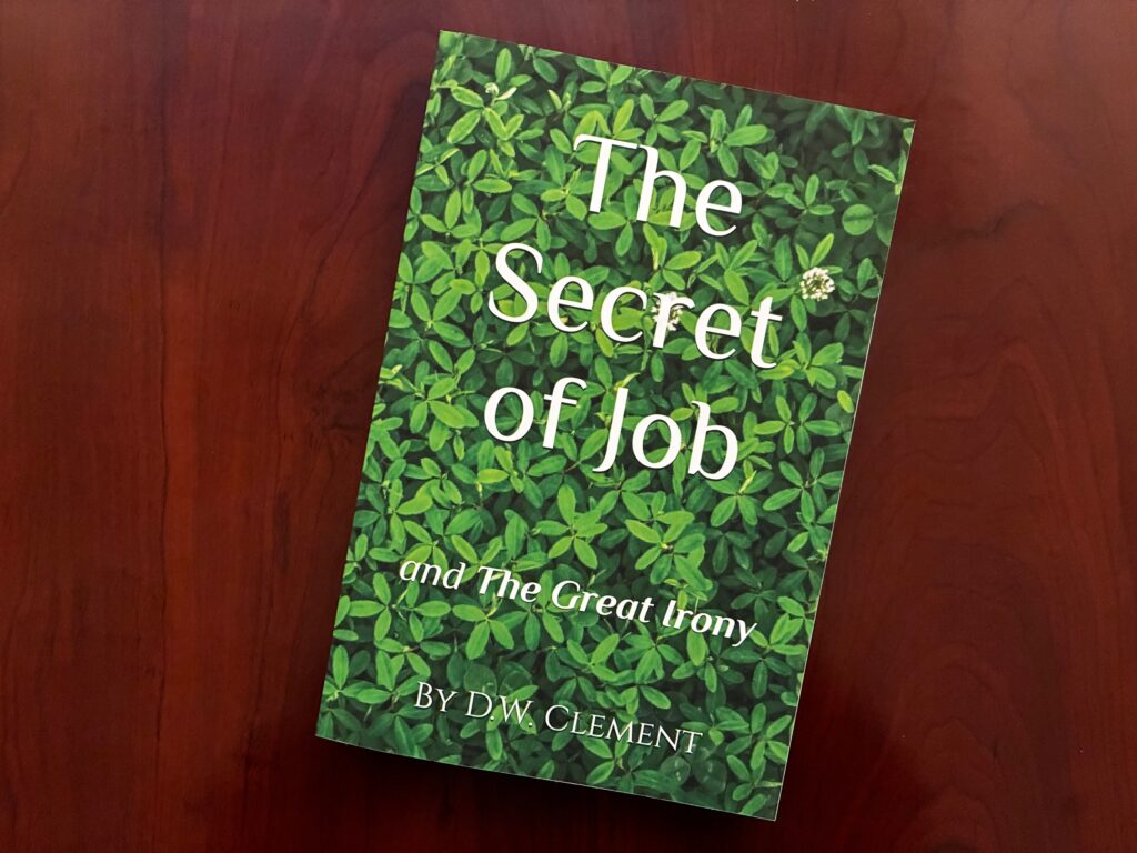 Book titled "The Secret of Job and The Great Irony" by D.W. Clement, featuring a green foliage background, resting on a wooden surface.
