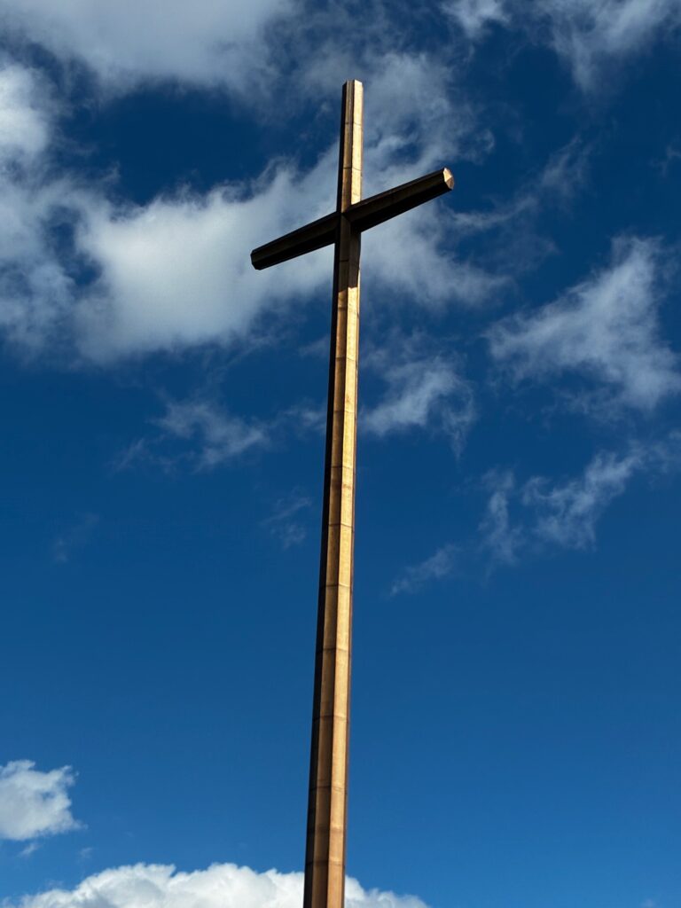 A tall wooden cross stands against a bright blue sky with wispy clouds, symbolizing faith and spirituality in a serene outdoor setting.