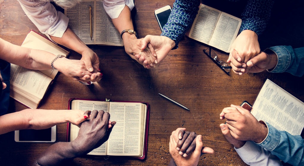 Diverse hands clasped together in prayer over open Bibles on a wooden table, symbolizing unity and faith during a group gathering.