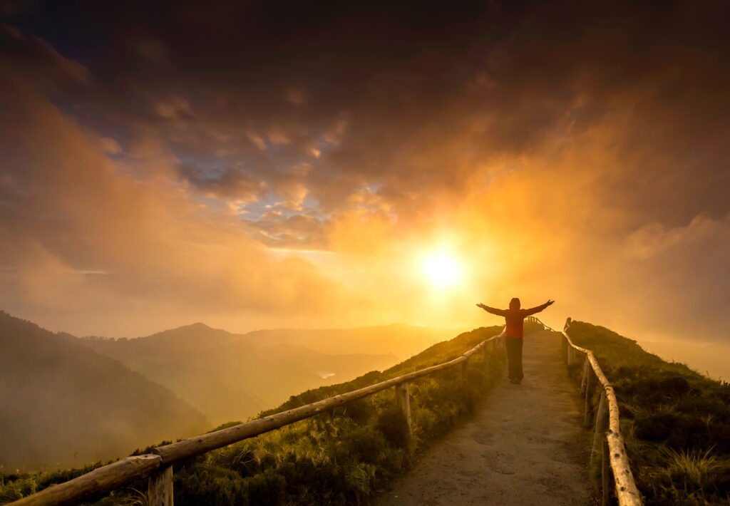 Silhouetted figure with outstretched arms stands on a path overlooking misty mountains at sunrise, symbolizing freedom and connection to nature.