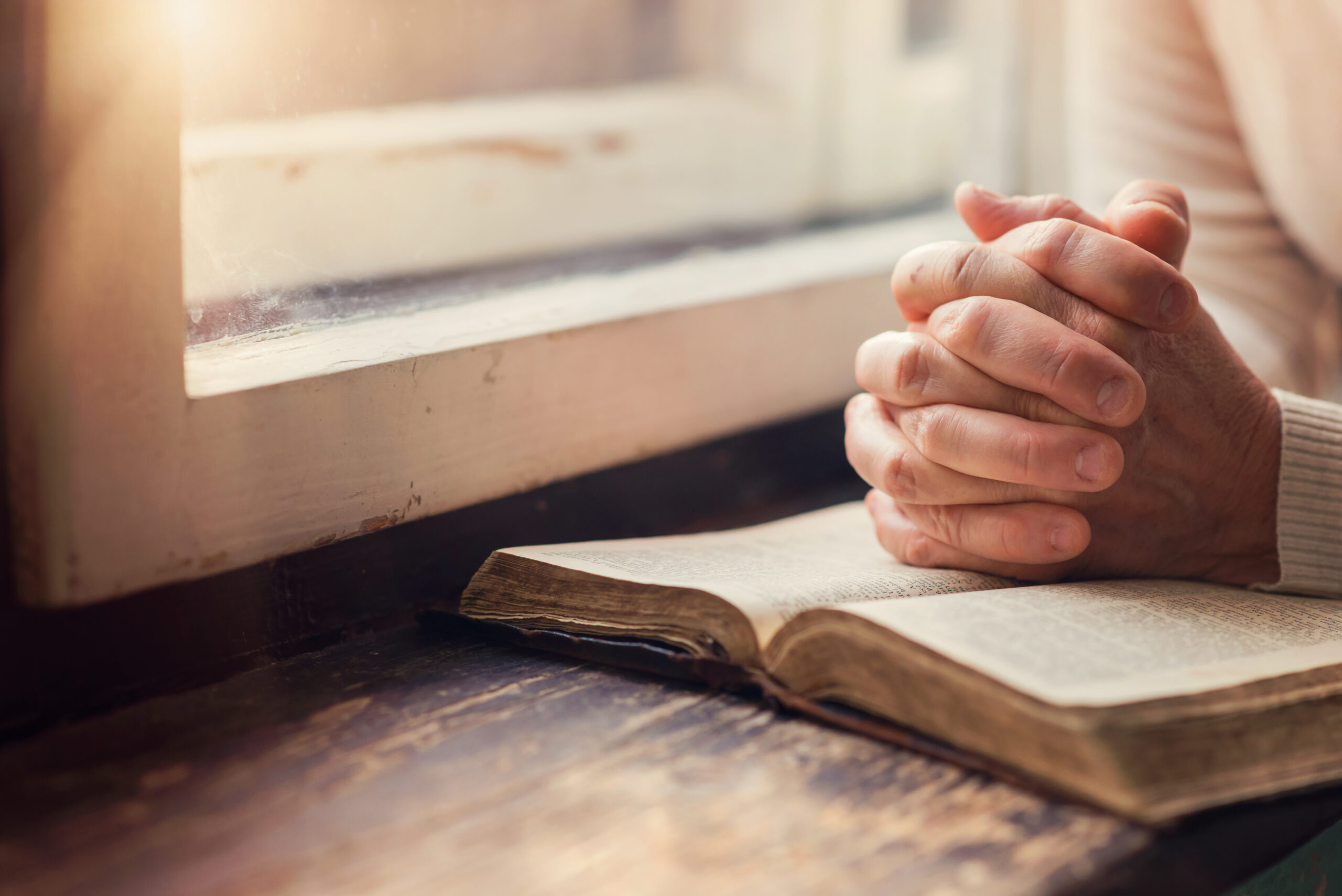 Hands are clasped in prayer over an open book on a wooden surface, with light streaming through a nearby window, evoking a sense of calm and contemplation.