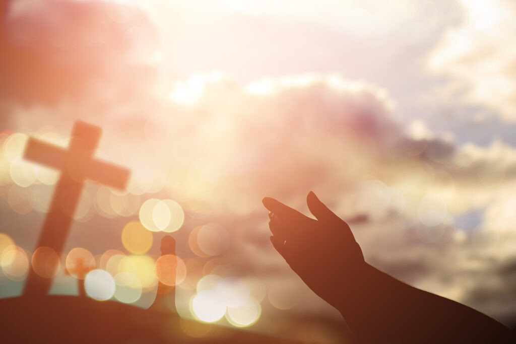 Hands reaching towards a cross against a glowing sunset, symbolizing hope and spirituality. The image captures a moment of prayer or reflection.