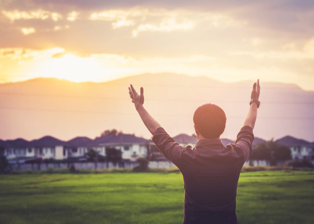 Person stands in a green field, arms raised towards a colorful sunset over distant mountains, symbolizing freedom and connection with nature.