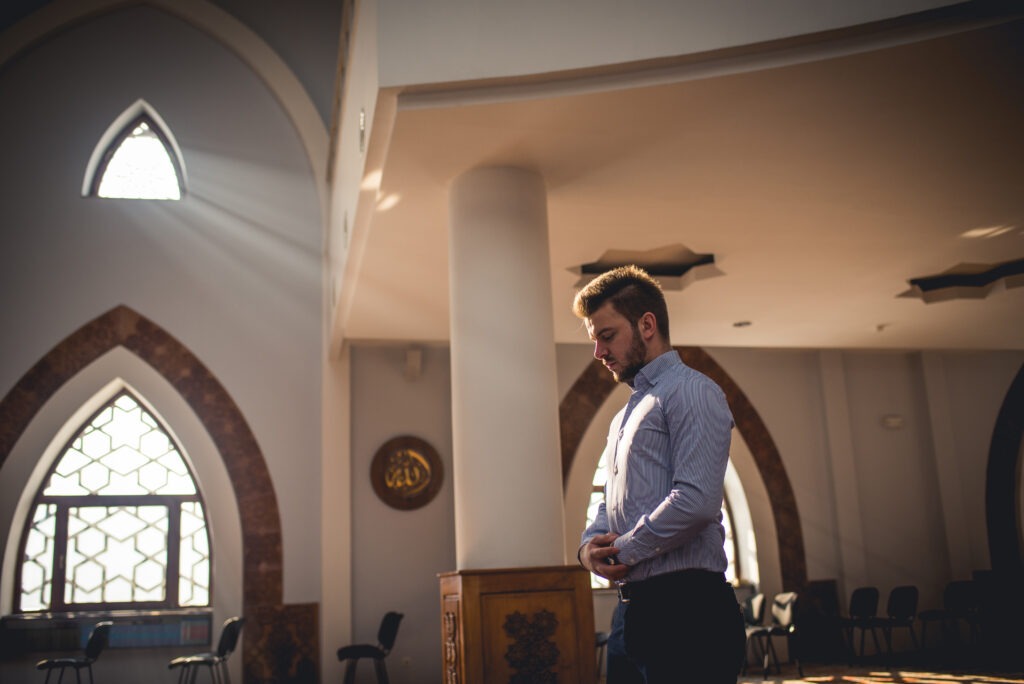 A young man stands in prayer within a sunlit mosque, framed by decorative arches, emphasizing a moment of spiritual reflection.