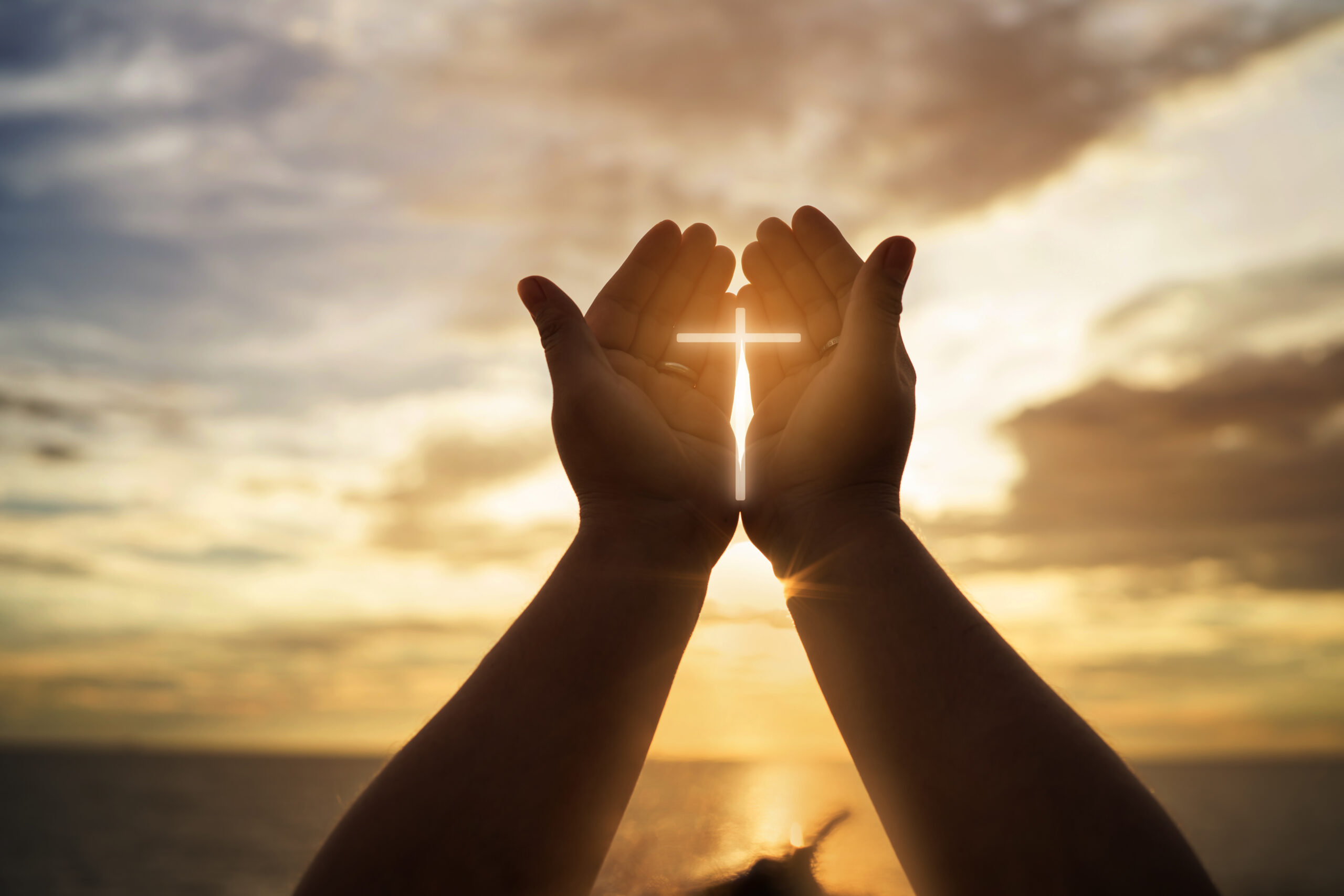 Hands uplifted against a sunset sky, holding a glowing cross, symbolizing faith and spiritual devotion.